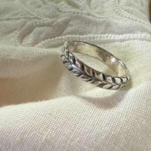 May include: A silver-toned metal ring featuring a continuous embossed leaf or wheat pattern around the entire outer band. The ring has a polished finish with darkened recessed areas that highlight the intricate, repeating botanical design.