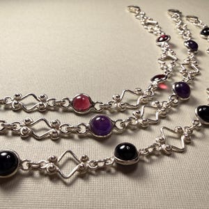 May include: Three silver-toned metal bracelets featuring a repeating pattern of open diamond-shaped links and circular settings. The bracelets are adorned with smooth, polished cabochon stones in shades of deep red, purple, and black.