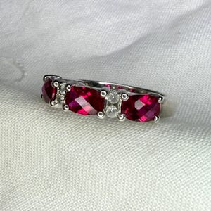 May include: A silver-toned ring featuring three cushion-cut vibrant red stones arranged in a row, separated by small, clear, round accent stones set in metal prongs. The band is smooth and metallic, showcasing a polished finish throughout the design.