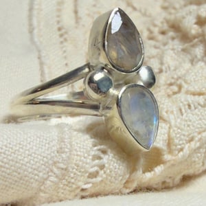 May include: A silver ring featuring a pear-shaped moonstone and two smaller round stones. The ring has a split shank design and is set on a cream-colored, textured surface. The moonstones have a subtle, iridescent glow.