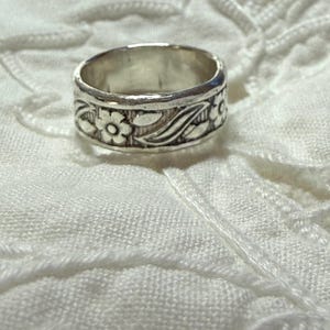 May include: A silver-toned metal ring featuring a continuous embossed pattern of small five-petaled flowers and elongated leaves encircling the entire outer band. The metal has a polished finish with darkened recessed areas to highlight the floral design.