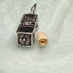 May include: Open silver-toned miniature box pendant with scroll. The box is square with ornate floral designs on the sides. The scroll is beige and rolled up. The pendant is on a light blue background.
