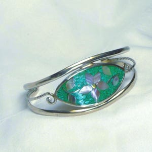 May include: Silver-tone cuff bracelet with an oval turquoise-colored centerpiece. The centerpiece features a flower design with purple petals and mother-of-pearl accents. The bracelet has a delicate, open design.