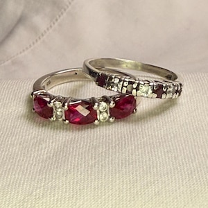May include: Two silver rings featuring vibrant red, faceted stones and small, clear accent stones. The front ring displays three larger red stones separated by pairs of clear stones, while the back ring features a row of alternating red and clear stones.
