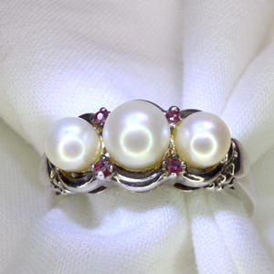 May include: A silver ring featuring three white pearls arranged in a row, with small red gemstones set between each pearl. The metal band has a polished finish and a curved design that follows the placement of the pearls and the red stones.