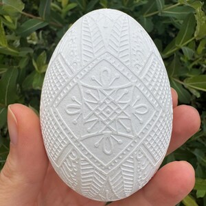 May include: A white, decorative egg with an intricate, embossed geometric pattern. The egg is held in a hand, with a green leafy background. The design features symmetrical shapes and lines, creating a detailed, textured surface.