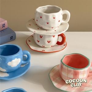 Könnte beinhalten: Eine Sammlung von Keramikbechern und Untertassen in verschiedenen Designs. Zwei weiße Becher mit roten Herzmustern sind gestapelt. Ein blauer Becher mit Wolkenmotiven und ein rosa Becher mit Karomuster sind ebenfalls zu sehen. Der Text "COCOON CLAY" befindet sich auf der rosa Untertasse.