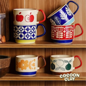 May include: A selection of ceramic mugs with diverse patterns, including apples, floral motifs, and geometric designs. The mugs display a range of colours, such as red, blue, yellow, and brown, with contrasting handle colours. The mugs are arranged on wooden shelves.