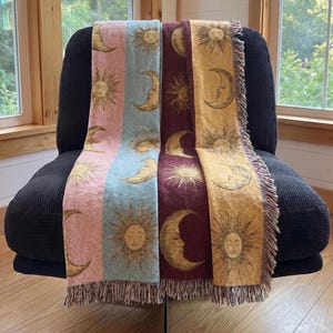 May include: A woven throw blanket featuring vertical panels in pastel pink, light blue, deep burgundy, and mustard yellow. The fabric is decorated with repeating gold-toned illustrations of suns and crescent moons with faces and fringed edges.