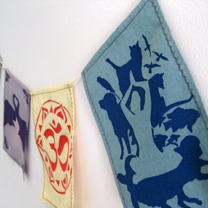 Könnte beinhalten: Eine farbenfrohe Stoffgirlande mit verschiedenen Designs. Die Girlande besteht aus rechteckigen Flaggen mit Bildern von Tieren, Symbolen und Mustern in Blau-, Lila-, Rosa- und Gelbtönen. Die Flaggen sind mit einem hellen Faden zusammengenäht.