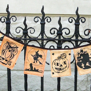 May include: A Halloween banner with orange fabric flags featuring black silhouettes of a cat, owl, witch, pumpkin, haunted house, and another cat. The banner is strung on a black metal fence.