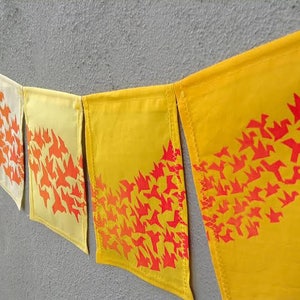 May include: A decorative banner with a series of fabric flags in shades of yellow and white. Each flag features a repeating pattern of orange bird silhouettes. The banner is hanging against a gray wall.