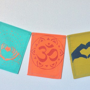 May include: A string of six colorful fabric flags with different symbols. The flags are pink, turquoise, orange, yellow, and pink. The symbols include a lotus flower, two hands forming a heart, the Om symbol, two hands forming a heart, and a heart with flames.