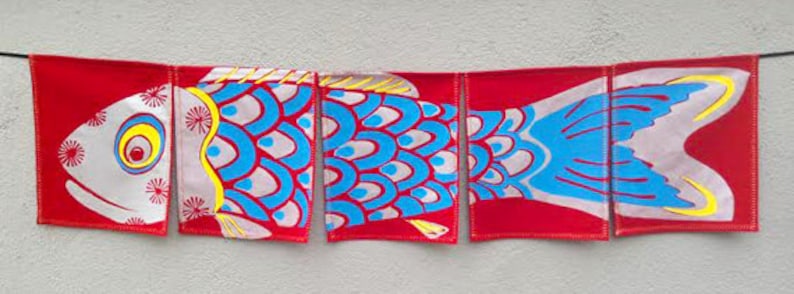 Puede incluir: Una pancarta colorida con forma de pez hecha de tela roja con detalles azules y blancos. La pancarta est&aacute; dividida en cinco secciones, cada una con una parte diferente del pez. El pez tiene un cuerpo blanco con escamas azules y una aleta amarilla.