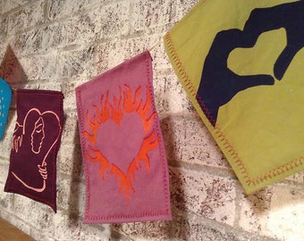 May include: Four fabric pennants with different designs. The first pennant is blue with a stylized image of a person. The second pennant is purple with a stylized image of two people embracing. The third pennant is pink with a stylized image of a heart with flames. The fourth pennant is yellow with a stylized image of two hands forming a heart shape.