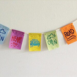 May include: A colorful banner with five small fabric flags strung together. Each flag is a different color: light blue, purple, yellow, light green, and orange. The flags have words and images like "OPEN", "HEART", a bus, and "ROAD".