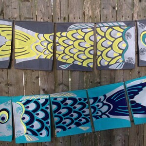 Puede incluir: Una hilera de siete banderas de tela con forma de pez, de colores vivos, colgadas de una valla de madera. Las banderas est&aacute;n hechas de tela gris claro con un dise&ntilde;o de pez azul y amarillo. Los peces tienen escamas y aletas, y algunos tienen ojos. Las banderas son una decoraci&oacute;n divertida y festiva para cualquier ocasi&oacute;n.