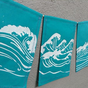 May include: A turquoise banner garland with a repeating white wave design. Each banner is rectangular and connected by a wooden dowel. The wave design is a stylized depiction of ocean waves.