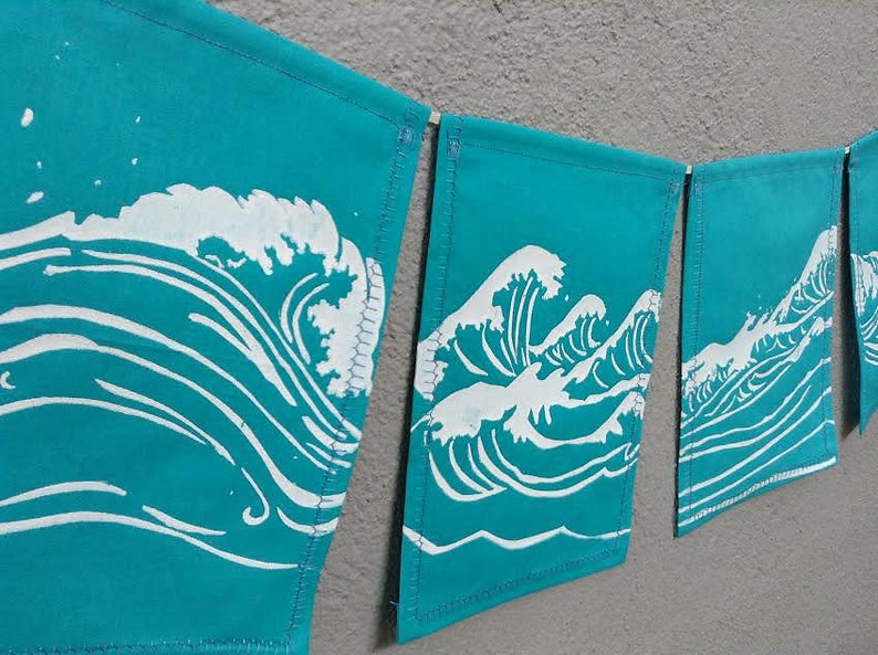May include: A turquoise banner garland with a repeating white wave design. Each banner is rectangular and connected by a wooden dowel. The wave design is a stylized depiction of ocean waves.
