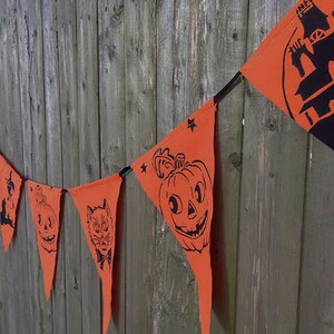 Puede incluir: Un bander&iacute;n de Halloween con banderines naranjas que presentan siluetas negras de calabazas, un gato, un fantasma y una casa encantada. El bander&iacute;n est&aacute; encordado en un cord&oacute;n negro y est&aacute; colgado en una valla de madera.