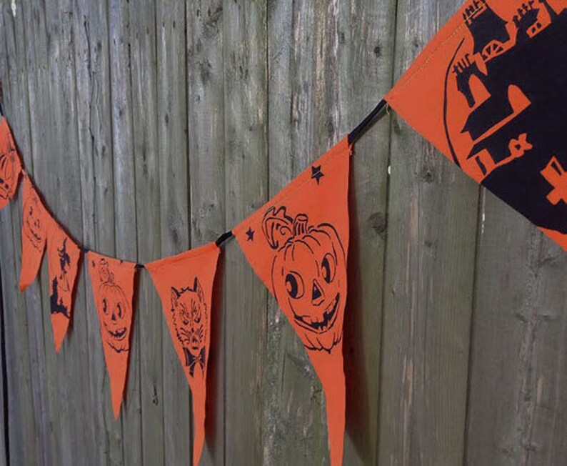 Puede incluir: Un bander&iacute;n de Halloween con banderines naranjas que presentan siluetas negras de calabazas, un gato, un fantasma y una casa encantada. El bander&iacute;n est&aacute; encordado en un cord&oacute;n negro y est&aacute; colgado en una valla de madera.