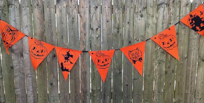 Puede incluir: Una pancarta de Halloween naranja y negra con varias im&aacute;genes, incluyendo una bruja, un gato, una calabaza y una casa encantada. La pancarta est&aacute; colgada en una valla de madera.
