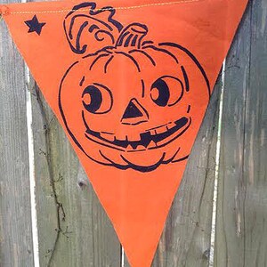 Puede incluir: Bandera de tela triangular naranja con una ilustraci&oacute;n en blanco y negro de una calabaza sonriente con ojos grandes.