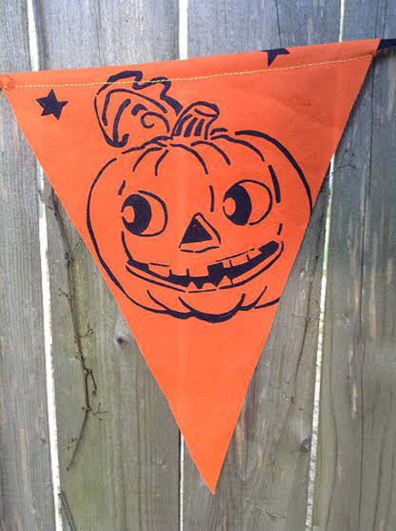 Puede incluir: Bandera de tela triangular naranja con una ilustraci&oacute;n en blanco y negro de una calabaza sonriente con ojos grandes.