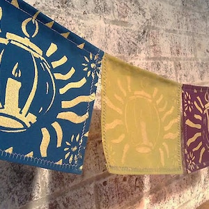 Pode incluir: Uma corda de bandeirinhas de tecido com um desenho de lanterna dourada sobre um fundo azul, amarelo, roxo e vermelho. As lanternas são contornadas em ouro e têm uma chama de vela dentro. As bandeirinhas estão penduradas em uma parede de tijolo.