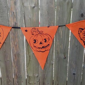 Puede incluir: Bandera de Halloween naranja con siluetas negras de una bruja volando en una escoba, una calabaza y un gato. La bandera est&aacute; colgada en una valla de madera.