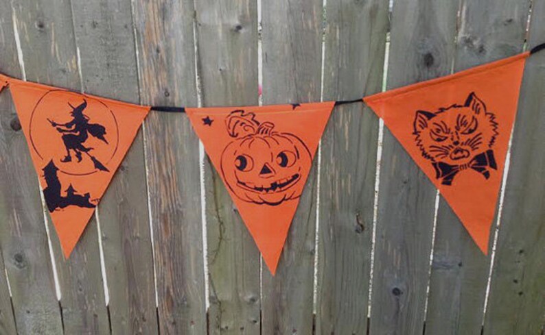 Puede incluir: Bandera de Halloween naranja con siluetas negras de una bruja volando en una escoba, una calabaza y un gato. La bandera est&aacute; colgada en una valla de madera.