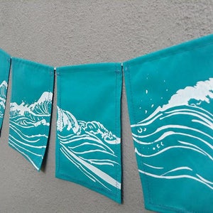 May include: A teal banner with white wave illustrations. The banner is made of rectangular flags strung together. Each flag features a different depiction of a wave, creating a cohesive ocean-themed decoration.