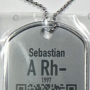 Könnte beinhalten: Silberfarbene Erkennungsmarke mit dem Namen "Sebastian", der Blutgruppe "A Rh-" und dem Jahr "1997" aufgedruckt. Ein QR-Code ist ebenfalls sichtbar. Die Marke ist an einer Kugelkette befestigt.