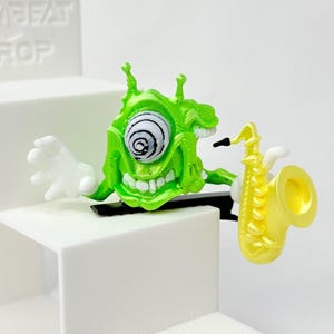 Puede incluir: Una figura de monstruo verde con un ojo grande y arremolinado y una amplia sonrisa, sosteniendo un saxofón amarillo. El monstruo tiene manos blancas y está posado como si estuviera tocando el instrumento. La figura está sobre una superficie blanca.