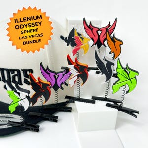 May include: A collection of colorful, stylized wing-shaped hair clips. Each clip features a unique color scheme, including red, orange, green, and purple. The clips are attached to black metal hair clips. A white hat with black text is in the background. The text "ILLENIUM ODYSSEY SPHERE LAS VEGAS BUNDLE" is on a sticker.