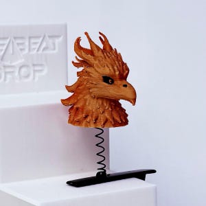 May include: A copper-colored 3D printed phoenix head figurine attached to a black metal spring and a black hair clip. The phoenix features textured, spiky plumage and dark, almond-shaped eyes. The object is mounted on a white tiered display stand.