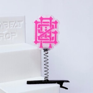 May include: A pink monogram hair clip with a black base and a silver spring. The monogram is a stylized design. The clip is on a white background.