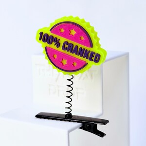 Peut inclure: Un badge circulaire jaune néon et rose vif avec le texte violet 100% CRANKED et quatre petites découpes en forme d'étoile. Le badge est fixé à un fin ressort hélicoïdal en métal noir monté sur une pince à cheveux noire de type crocodile.