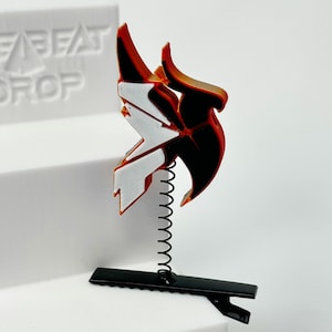 May include: A red, black, and white decorative object with a spring and clip base. The object has a stylized, angular design. The base is black, and the spring is silver. The object is displayed on a white surface.