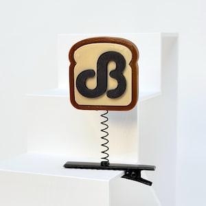 May include: A hair clip featuring a slice of toast with a brown crust and a tan center. A black letter B is centered on the toast. The toast is attached to a black metal hair clip by a thin, coiled black metal spring that allows for movement.