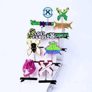 May include: A collection of ten hair clips featuring various designs including the text Excision, Lost Lands, and Shrek, along with a pink dinosaur skull, a black skull on an X, and green X shapes, all mounted on metal springs and black clips.