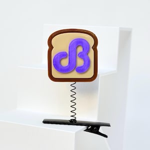 May include: A toast-shaped badge with a brown crust and tan center, featuring a raised purple letter B in a stylized font. The badge is attached to a thin black metal coil spring, which is connected to a black alligator-style hair clip.