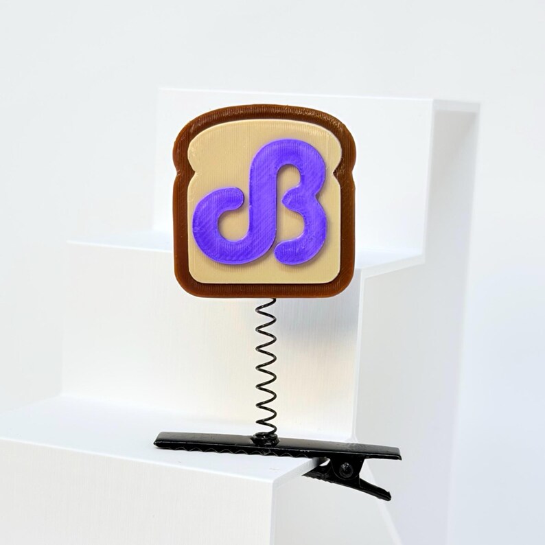May include: A toast-shaped badge with a brown crust and tan center, featuring a raised purple letter B in a stylized font. The badge is attached to a thin black metal coil spring, which is connected to a black alligator-style hair clip.
