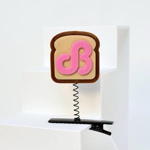 May include: A hair clip featuring a brown and tan toast-shaped topper with a pink stylized letter B on the front. The toast is attached to a thin black metal spring, which is connected to a black alligator-style hair clip base for attachment.