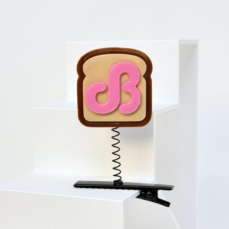 May include: A hair clip featuring a brown and tan toast-shaped topper with a pink stylized letter B on the front. The toast is attached to a thin black metal spring, which is connected to a black alligator-style hair clip base for attachment.