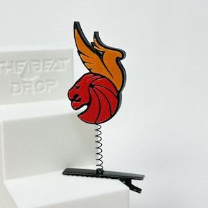 May include: A decorative hair clip featuring a red lion with orange wings and a black outline. The clip is attached to a black base with a spring and a clip. The text "THE BEAT DROP" is visible in the background.