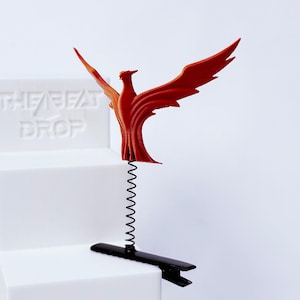 May include: A red phoenix figurine with outstretched wings attached to a thin black metal spring and a black alligator hair clip. The phoenix has a textured surface and is positioned above a white block with the embossed text THE BEAT DROP.