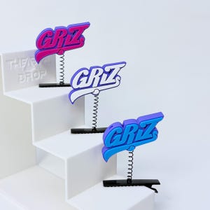 May include: Three hair clips featuring the text GRIZ in different colour combinations. Each clip has a black metal base, a thin black spring, and a raised plastic logo in purple and pink, white and purple, or purple and blue colour schemes.