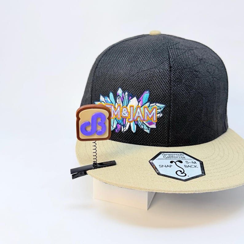 May include: A black baseball cap with a tan brim, featuring an embroidered graphic of crystals and the text "EM & JAM". A brown toast-shaped pin with a purple "JB" logo is attached to the brim by a black clip and a small metal spring.