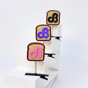 May include: Three hair clips featuring a slice of toast design with a brown crust and tan center. Each toast has a raised letter B in black, purple, or pink. The toast is attached to a black metal alligator clip by a thin, flexible metal spring.
